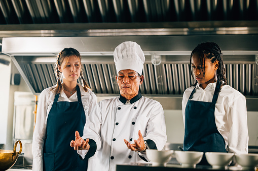 Professional Asian senior chef guides diverse students in a restaurant kitchen workshop focusing on teamwork learning and note-taking. Educational setting. Food Edocation