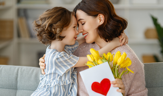 Happy mother's day. Child daughter congratulating mom and giving her flowers and postcard. Mum and girl smiling and hugging. Family holiday and togetherness.