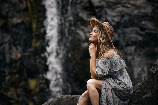 Smiling woman with a hat day dreaming while sitting on the rock by the waterfall in nature.