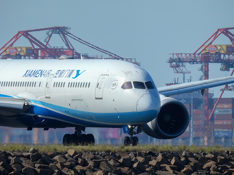 A Xiamen Air Boeing B787-9 plane, registration B-7836, taxiing after arrival at Sydney Kingsford-Smith Airport as flight MF801 from Xiamen. Heat from the engines creates a heat haze under the plane.  In the background is a container ship and cranes at the Port Botany Container Terminal. This image was taken from near Kyeemagh Beach, Botany Bay on a hot and sunny morning on 24 March 2024.
