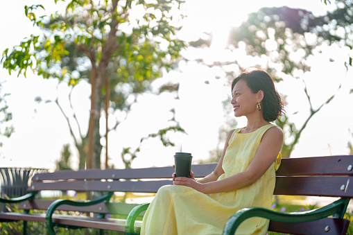 Portrait of a young Asian woman enjoying a cup of coffee in the park, savoring a beautiful day outdoors and breathing in the fresh air.