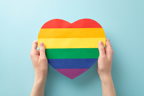A first person top-down view of a woman's hands holding a rainbow-colored heart on a pastel blue backdrop with an open area for text or advertisement, alluding to LGBT History Month's theme