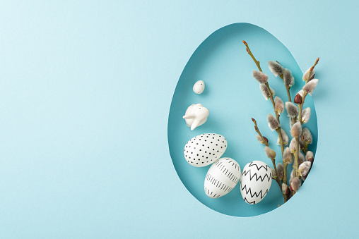 Joyful Easter inspiration: Shot from top view capturing winsome Easter Hare sculpture, accompanied by soft pussy willow and eggs, through egg-like gap on pastel blue surface, blank space for greetings
