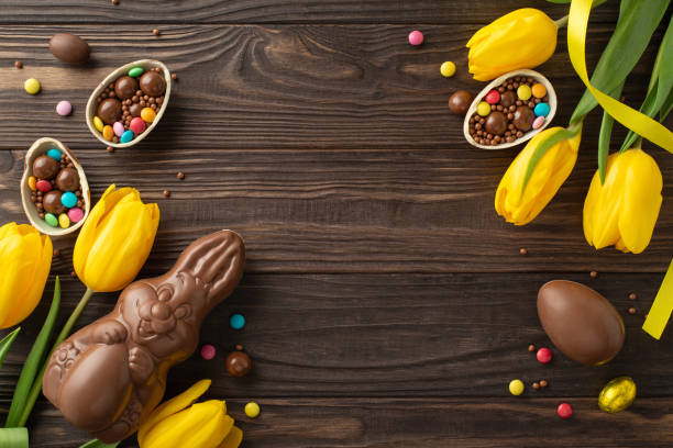 istock Easter candy scenario: Top view shot of chocolate eggs split open with colorful candy contents, a chocolate bunny, and tulips against a wood setting, space reserved for text or advert insertion 2111423757
