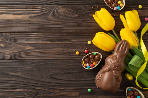istock Charming Easter sweets setup: View from top of chocolate eggs cracked to reveal a rainbow of candies, with a chocolate bunny and bunch of tulips on a wooden backdrop, space for text or ads 2111423758