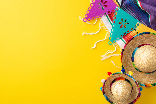 Cinco de Mayo theme. Overhead shot featuring festive items: mini sombreros, colorful serape, string of flags on a vibrant yellow backdrop, space for text