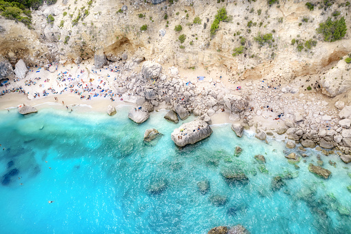 Summer holiday background. Aerial view of seascape with stone coastline and small beach with crowd of people. Sea coast with blue, turquoise clear water. Islands of Sardinia in Italy