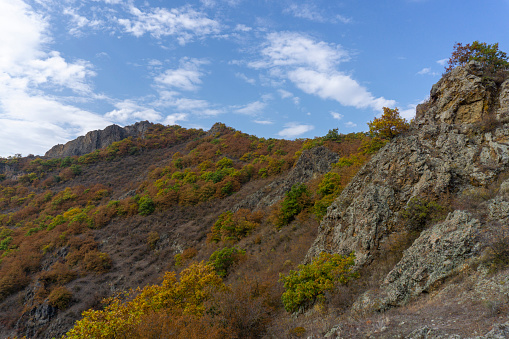 View of the mountains and rocks from the hill. Autumn colors of red, yellow, green and orange leaves and bushes. Clear blue sky and clouds. Hills and peaks.