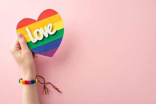 First person top view photo of young woman's hand with symbolic bracelet holding rainbow heart card with text love on pastel pink background with empty space for text or advert