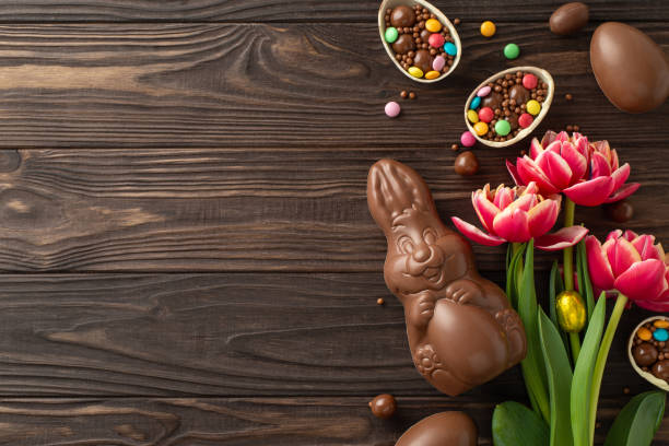 istock Sweet Easter fantasy composition. Top view perspective of chocolate eggs opened to show colorful candy treasures, a cocoa bunny, and spring tulips on a wood backdrop, with area for copy or adverts 2111492848