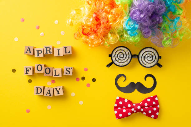 istock Prank top view setup, showcasing 'April Fool Day' spelt on cubes, alongside whimsical items like clown wig, specs, bow tie, mustaches, confetti, all arrayed to mimic festive face on yellow backdrop 2111492849