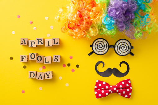 Prank top view setup, showcasing 'April Fool Day' spelt on cubes, alongside whimsical items like clown wig, specs, bow tie, mustaches, confetti, all arrayed to mimic festive face on yellow backdrop