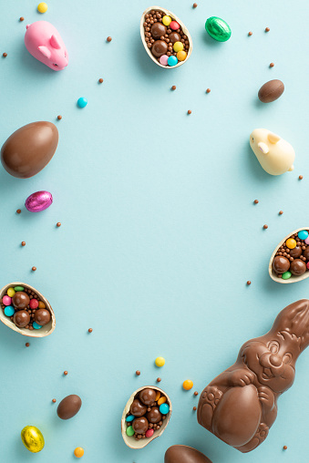 Lovely Easter collection theme. Top view vertical photo of fragmented chocolate eggs, overflowing with colorful candies, chocolate bunny, sprinkles on light blue backdrop, with area for text or advert