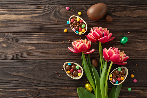 Festive Easter presentation. View from top of shattered chocolate eggs filled with colorful confections, and fresh tulips arranged on a wooden table, with blank space for messages or advertising