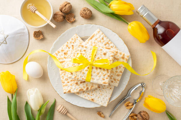 istock Seder night layout idea: Overhead shot featuring ribbon-tied matzo, crimson wine, goblet, walnuts, boiled egg, honey and spoon, nutcracker, kippah plus tulips against a soft beige linen backdrop 2111492860