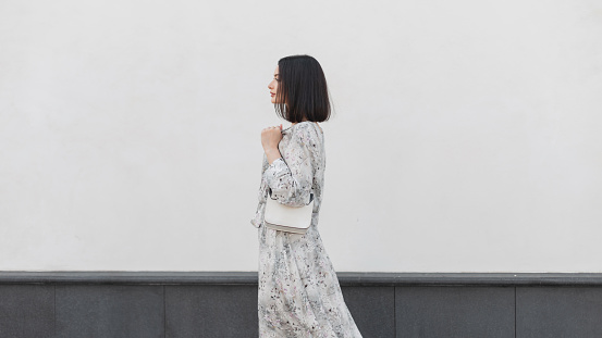 Beautiful elegant fashion girl with hairstyle in a fashionable flowers dress with a stylish bag walks near a white wall in the city, side view