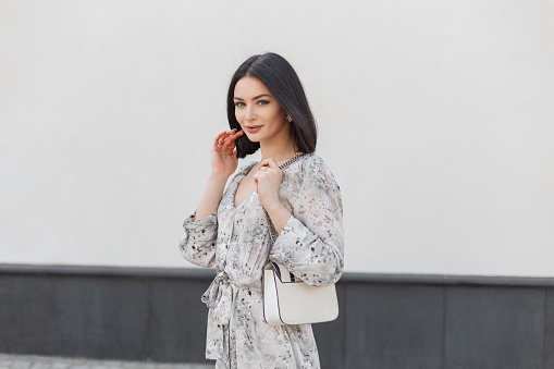 Fashionable pretty brunette woman model with shorts hairstyle in stylish vintage flowers dress with fashion white bag walks near a white building on the street