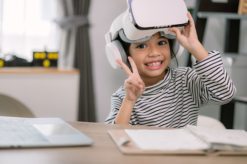 Cute Asian little girl wearing VR glasses with a laptop placed on the table in STEM technology class. Online education. Erudition.