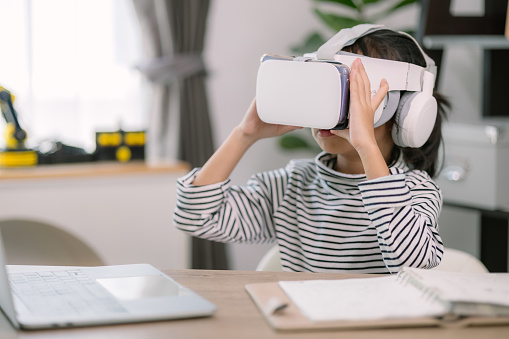 Cute Asian little girl wearing VR glasses with a laptop placed on the table in STEM technology class. Online education. Erudition.