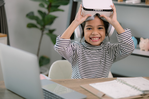 Cute Asian little girl wearing VR glasses with a laptop placed on the table in STEM technology class. Online education. Erudition.