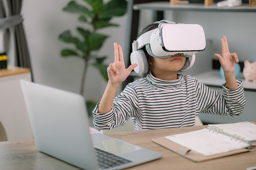 Cute Asian little girl wearing VR glasses with a laptop placed on the table in STEM technology class. Online education. Erudition.