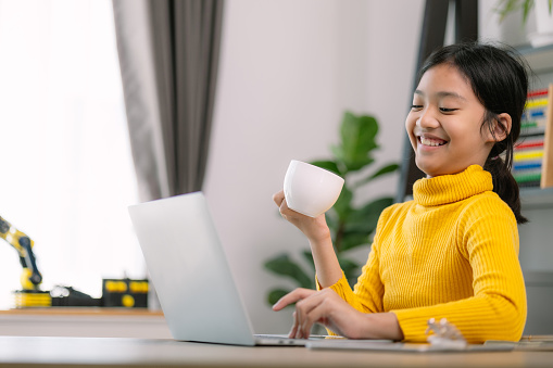 A young girl is sitting at a desk with a laptop and a cup of coffee. She is smiling and she is enjoying her time