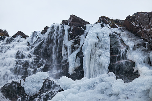 Partially frozen waterfall flows amidst ice formations on a rugged cliffside during a cold winter day