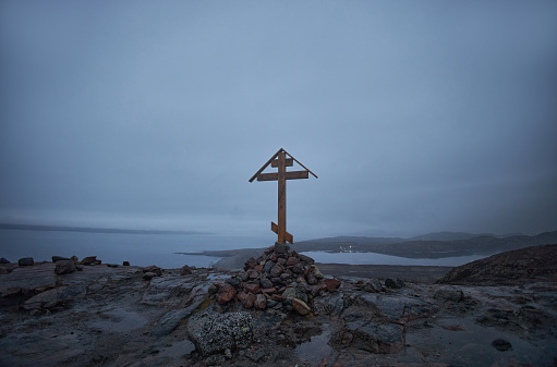 Simple wooden cross atop a rocky hill, with a foggy coastal backdrop under the evening sky