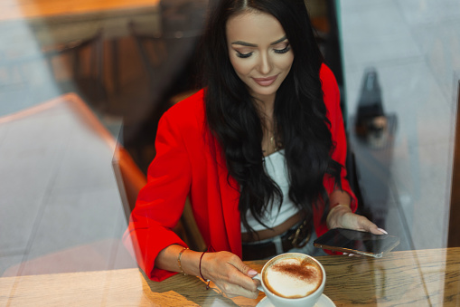 Beautiful successful businesswoman in fashion bright clothes with a red blazer is sitting in a cafe with a smartphone, drinking coffee. Girl rest in cafe behind glass with reflection