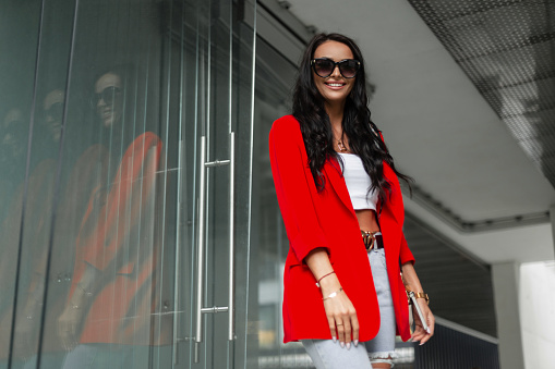 Happiness young beautiful stylish woman with smile with trendy sunglasses in fashion casual business outfit with red blazer, top and jeans walks on the street on a modern urban background