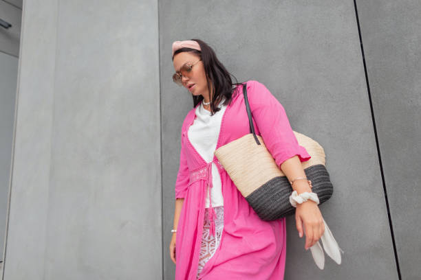 fashionable beautiful girl model with cool sunglasses in trendy summer clothes with a pink pareo, white tank top with a wicker straw beach bag near a gray concrete wall on the street - women sarong beach white стоковые фото и изображения