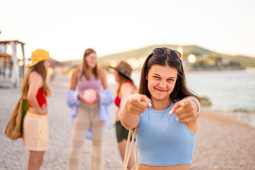 Portrait of a young teenage girl with long dark hair, wearing a light blue tank top and sunglasses on the top of the head and pointing at camera with both hands. Group of young girls in the blurred background on the beach early in the morning.