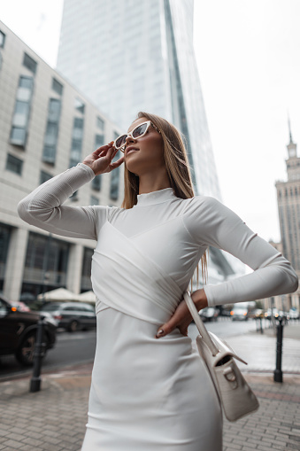 Fashion beautiful young woman in a white fashion dress with a handbag puts on white stylish sunglasses and walks in the city. Urban fashion vogue and beauty