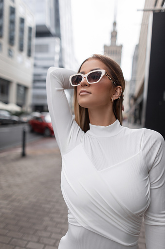 Outdoor female portrait of a stylish beautiful vogue woman with white cool sunglasses in a fashiona white dress walks in the city