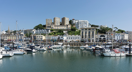 View across Torquay harbour in Devon, UK.  Hotels and apartments can be seen in the distance and boats can be seen moored in the harbour.  People can be seen on the promenade.