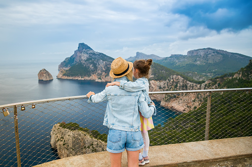 A woman in straw hat hugging her little daughter, while gazing at a breathtaking view from Mirador de El Colomer in Mallorca. Travelling with children concept.