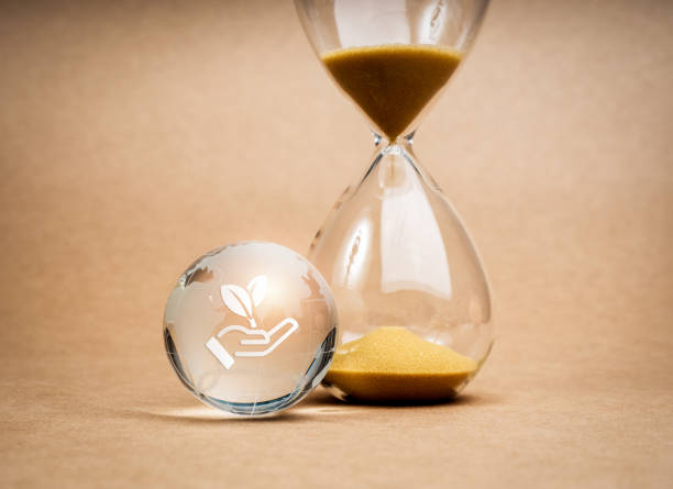 istock Environmental sustainability, time and responsibility, conserve resources concept. The environment care icon, hand holding leaf, on small glass world globe near hourglass on recycle paper background. 2111678680