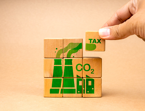 CO2, carbon TAX, global greenhouse gas emissions. Text TAX piece block in hand put on cube puzzle stack wood blocks with green factory icon. Environmental and social responsibility business concept.