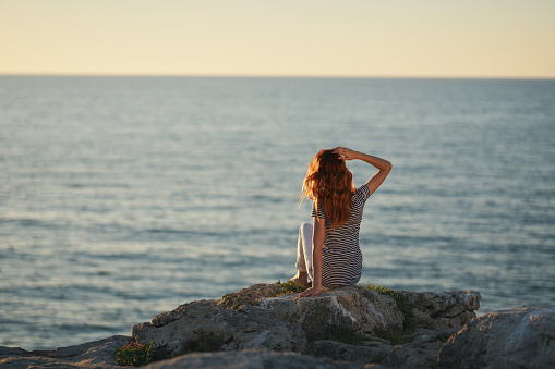 a woman sits on a Big Stone near the sea and looks at the sunset in the mountains. High quality photo