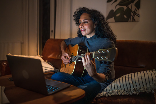 Smiling woman learning to play guitar of online lesson on laptop at home.