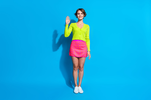 Full body length photo of beautiful lady wearing green cardigan and mini skirt waving arm to say hi isolated on aquamarine color background.