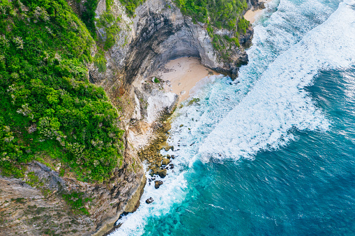 Secluded beach cove with golden sands surrounded by rocky cliffs covered in lush greenery. Waves from the turquoise ocean crash against the rocks and shore, showcasing nature’s raw beauty. Shot taken in Bali, Bukit.