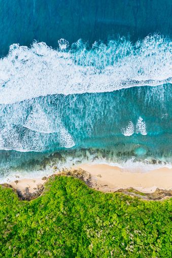 Aerial view of waves crashing onto a sandy beach, creating white foam. The turquoise ocean contrasts with the beige coastline. Lush green vegetation borders the shore, framing this natural coastal scene. Shot taken in Bali, Bukit.