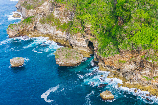 Cliff adorned with greenery overlooks a turquoise sea, waves crashing against the rocky shore. Natural rock formations protrude from the water, showcasing the raw beauty of this coastal landscape. Shot taken in Bali, Bukit.