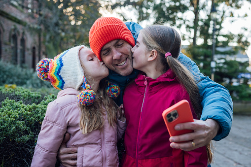 A medium close-up of a father taking a selfie with his daughters along the street of South Shields, North East England. The two daughters kiss the father's cheeks whilst he smiles happily. They are all wearing casual winter clothing.