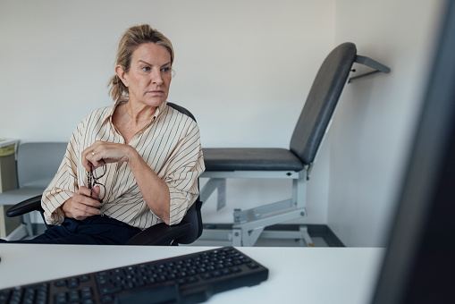 Female general practitioner sitting at her desk with a computer. She is looking at her computer screen with a contemplative expression.