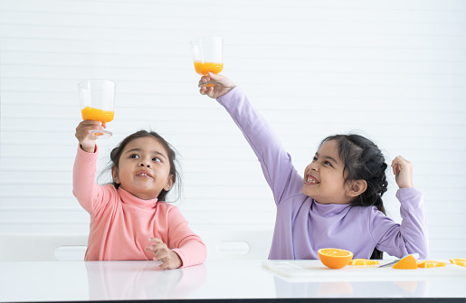 Cheerful Asian little children girls holding, drinking fresh orange juice from glass and raising glass up after making orange juice together at home. Nutrition, Healthy food. White background