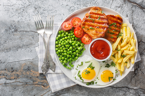 Fried pork loin steak served with French fries, fied eggs, green pea and fresh tomato closeup on the plate on the table. Horizontal top view from above