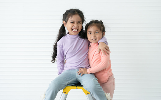 Portrait of Asian two little sisters, 6 year and 3 year old, in casual clothes with black long hairs. Sibling laughing sitting and standing on a ladder, embracing each other and looking at camera