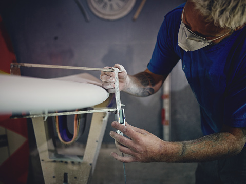 Crop attentive male shaper with blond hair and tattoos on arms in casual clothes and respirator measuring surfboard thickness with caliper during work in joinery